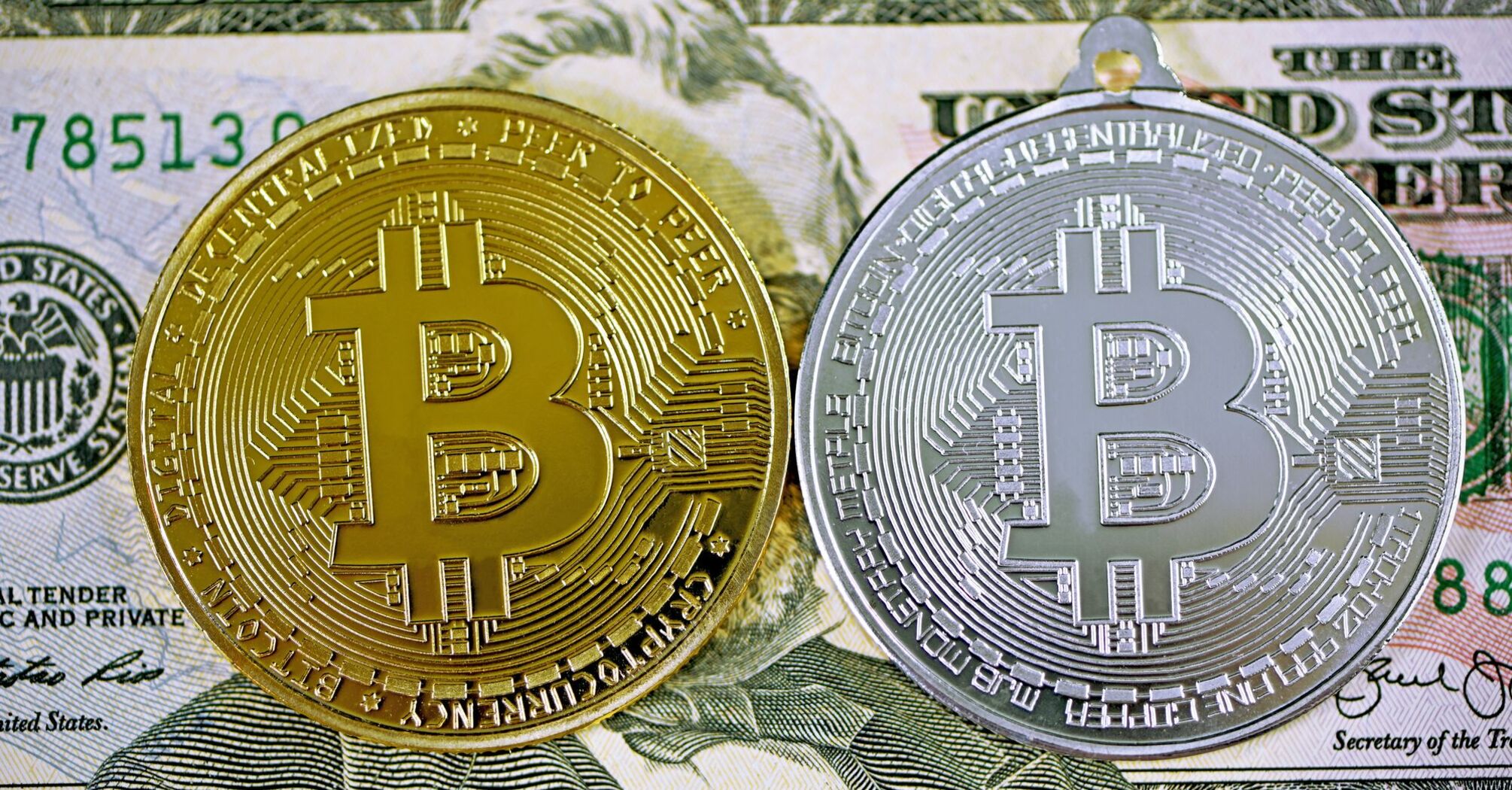 Дві монети Bitcoin – золота та срібна – лежать на банкноті 100 доларів США